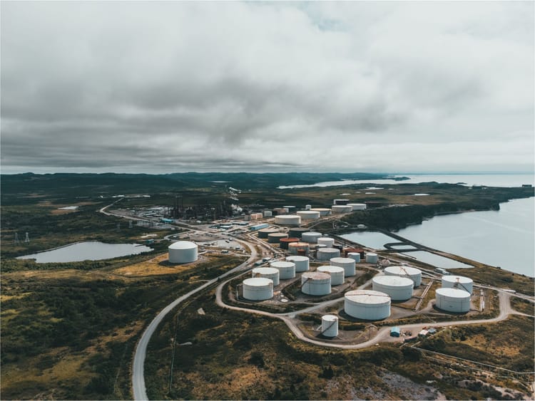 Aerial view of coastal oil storage tanks and refinery infrastructure beside a body of water.